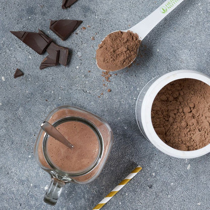 Chocolate powder in a spoon, jar, and cup on a gray surface with chocolate pieces.
